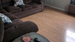 A living room with a brown sofa, a small table with remotes, and a lamp on a side table. - Cozy camp located in Elk County adjoining state game land. (Byrnedale)