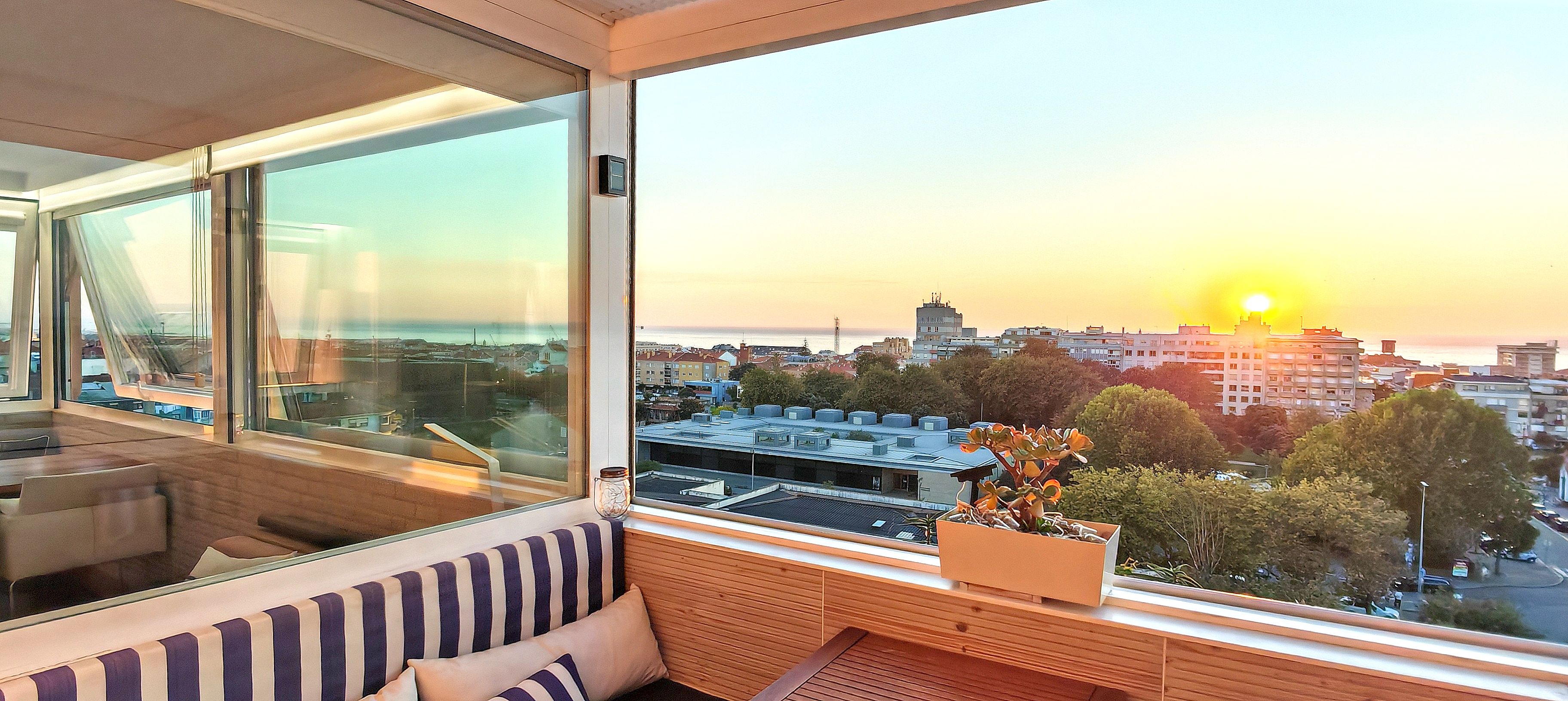 A room with a large window overlooking a cityscape at sunset, a wooden bench, and a potted plant.