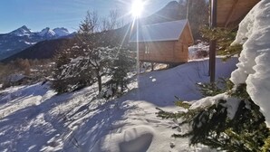 Een houten chalet met een schuin dak, omgeven door met sneeuw bedekte grond en bomen, met bergen op de achtergrond.