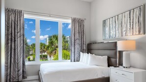 A modern bedroom with a large bed, a bedside table with a lamp, and a view of palm trees through the window.