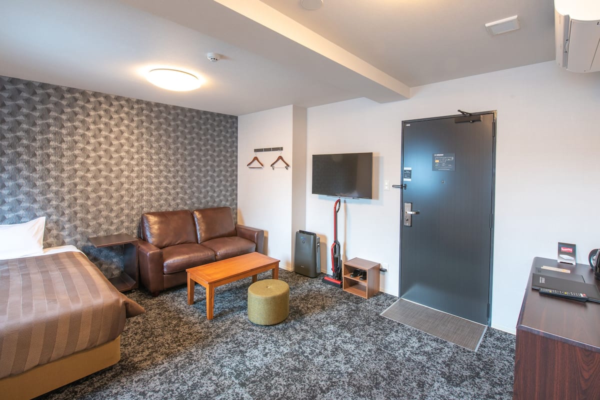 Quarto de hotel com cama, sofá, mesa pequena, TV presa na parede e porta com aviso.