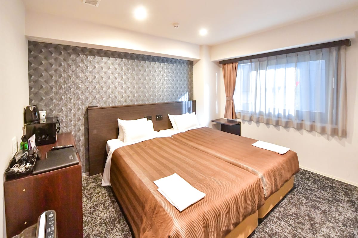 Quarto de hotel com uma cama grande, uma escrivaninha com um laptop, uma máquina de café e uma janela com cortinas.