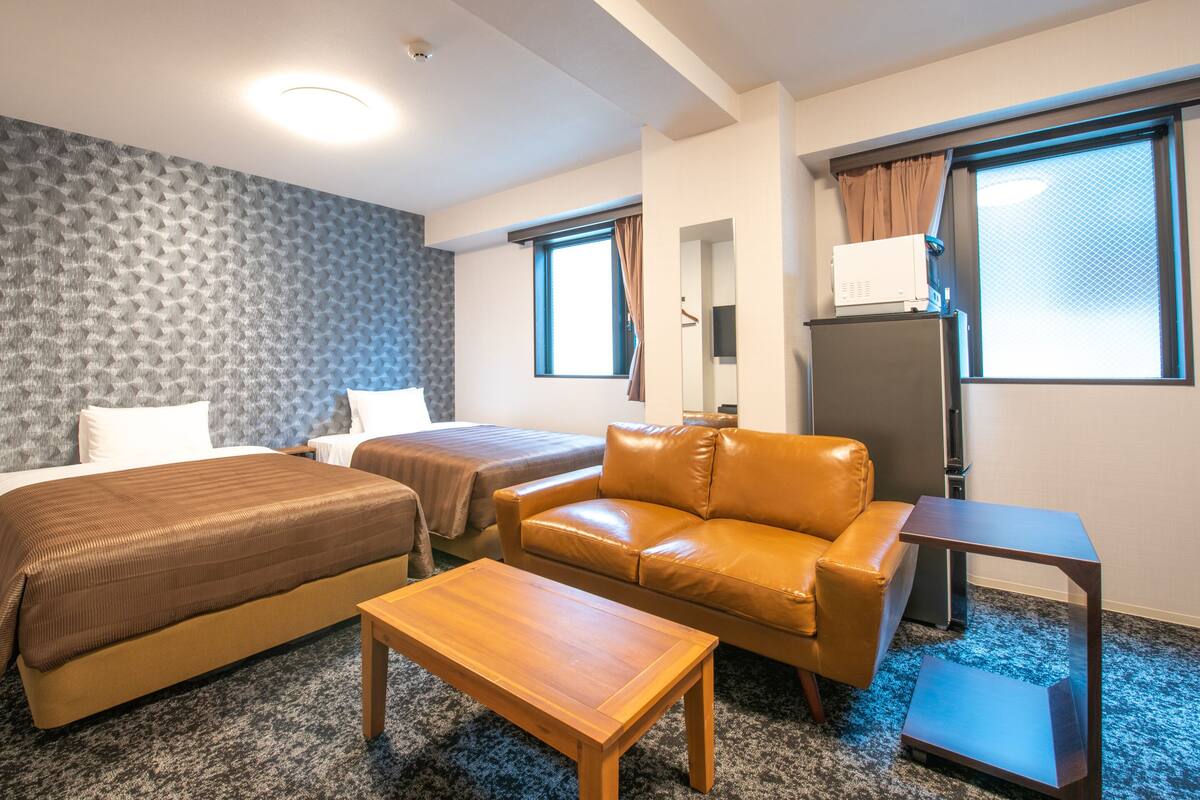 Quarto de hotel com duas camas, um sofá de couro, uma mesa de centro de madeira, uma mesa pequena e um refrigerador.