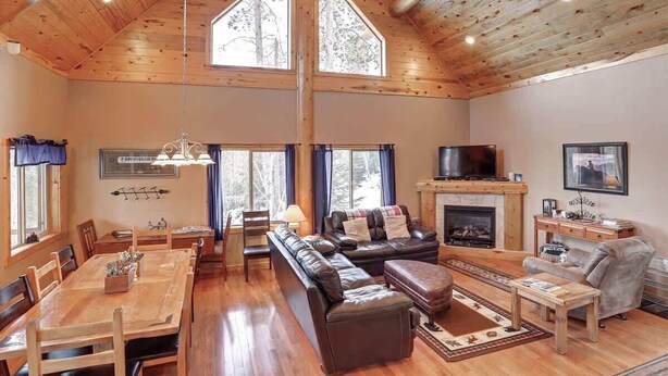 A spacious living room with a high ceiling, wooden beams, a fireplace, a television, and a dining area with a table and chairs. - Arrow Lodge (Lead)