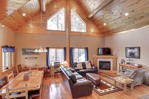 A spacious living room with a high ceiling, wooden beams, a fireplace, a television, and a dining area with a table and chairs. - Arrow Lodge (Lead)