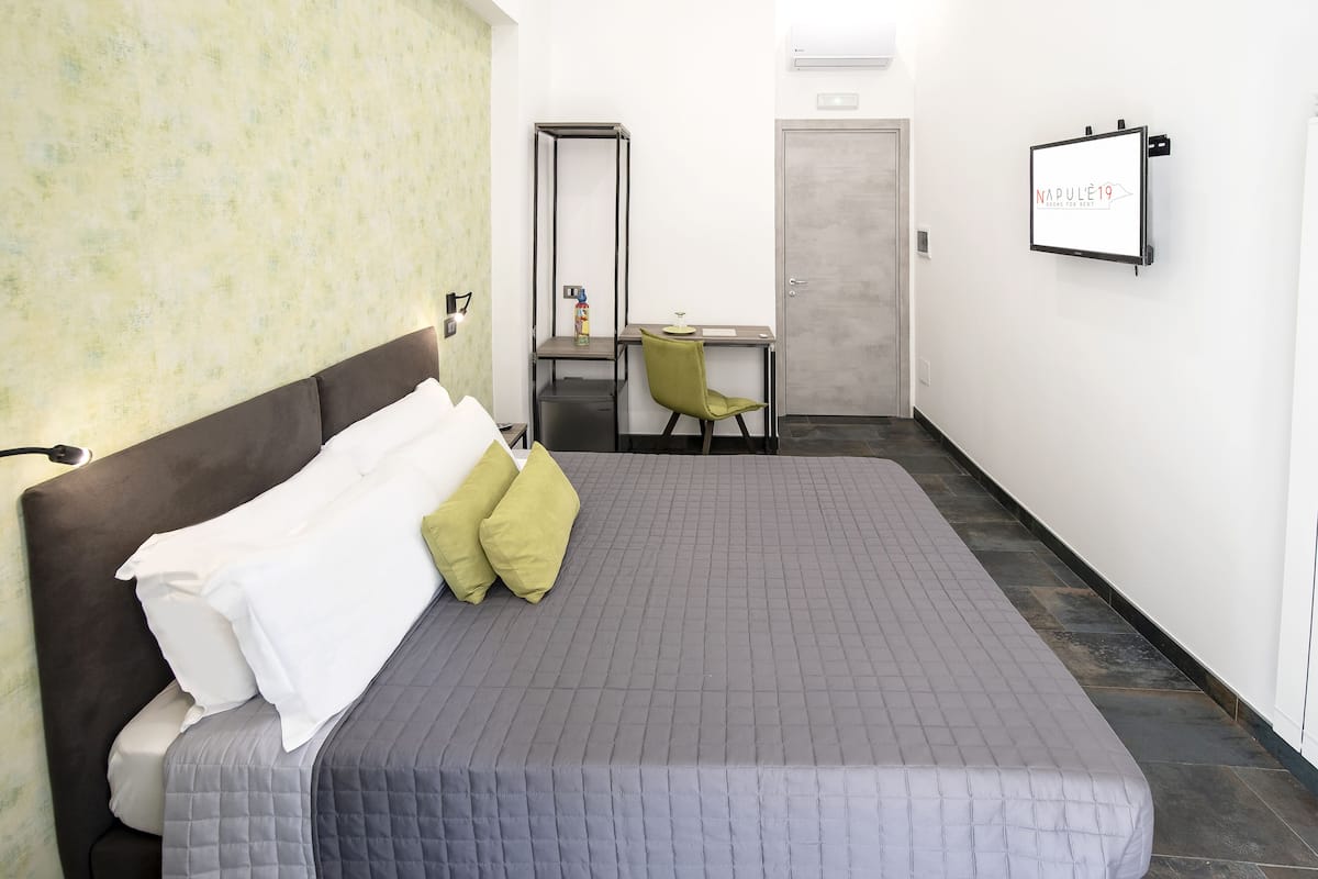 Habitación de hotel con cama, escritorio, silla, armario y una pantalla de televisión mostrando 'NAPOLLE19'.
