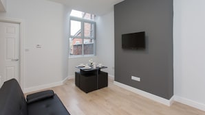 Una sala de estar moderna con un sofá negro, una mesita baja con tazas, un televisor montado en una pared gris y una ventana con vistas a edificios.