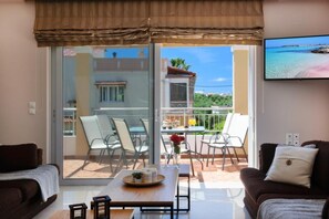 A living room with a sofa, a coffee table, and a TV mounted on the wall. There is a balcony with chairs and a table, and a view of a building and greenery.
