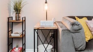 A modern living room with a grey sofa, a small wooden side table with a lamp, a white bucket, and a plant on a black metal shelf.