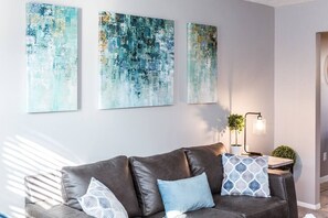 Una sala de estar moderna con un sofá de cuero, tres cuadros abstractos, una mesita con una lámpara y una planta en maceta.