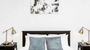 Ein ordentlich gemachtes Bett mit zwei Kissen, einem dunklen Kopfteil und einer weißen Backsteinwand. An beiden Seiten des Bettes steht jeweils ein schwarzer Nachttisch mit einer Lampe und einer kleinen Pflanze.