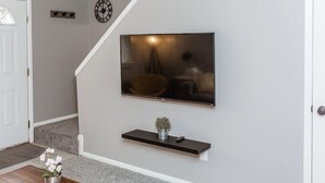 A modern living room with a flat-screen TV mounted on the wall, a small shelf below it, and a dining table with a flower vase.