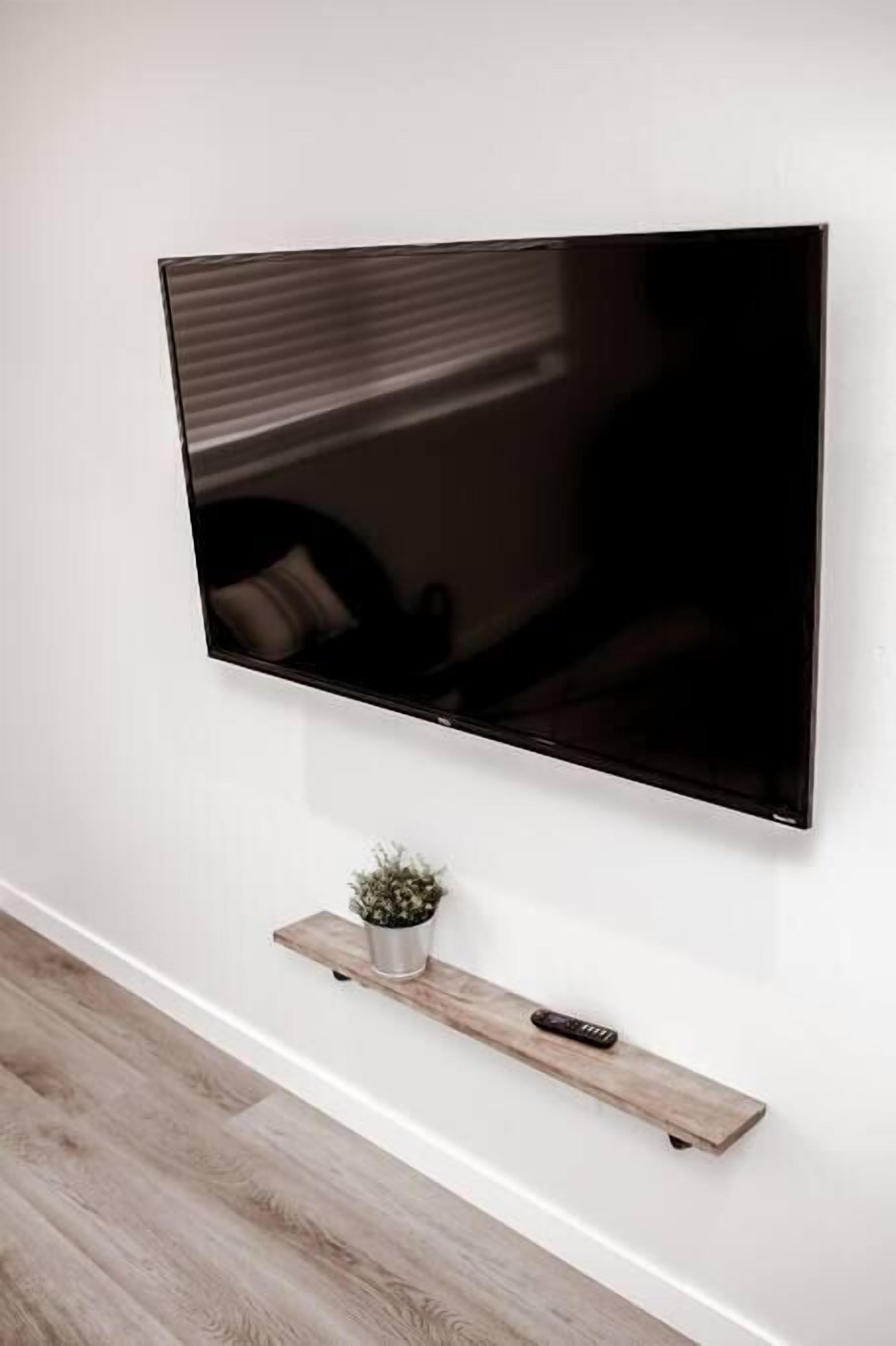 A flat-screen TV mounted on a white wall, with a wooden shelf below holding a small potted plant and a remote control.