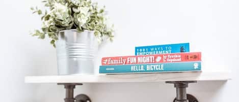 A white shelf with a metal pot containing green plants, and three books titled '1001 WAYS TO EMPOWERMENT', 'family FUN NIGHT!', and 'HELLO, BICYCLE'.