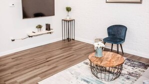 A modern living room with a wooden floor, a round coffee table with a vase of flowers, a blue chair, a TV mounted on the wall, and a small shelf with decorative items. - Renovated 2story (9C) OSU/Central ❤Near EVERYTHING (Columbus)