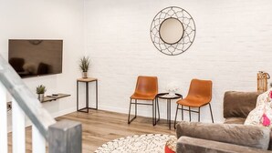 Una sala de estar moderna con una pared de ladrillo blanco, un espejo redondo, un televisor de pantalla plana, dos sillas naranjas, un sofá marrón y una pequeña mesa con una planta en maceta.