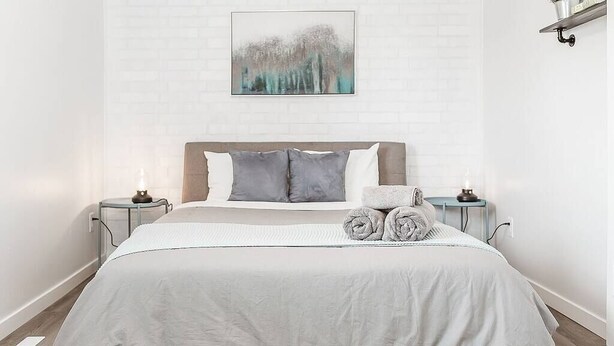 Una cama bien hecha con edredón gris y blanco, dos almohadas grises y toallas enrolladas en la barra de la cama.