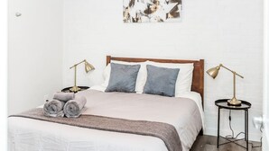 Un lit bien fait, avec une couette blanche et deux oreillers, un plaid marron et deux serviettes enroulées posées sur le côté. Il y a deux tables de chevet avec des lampes.