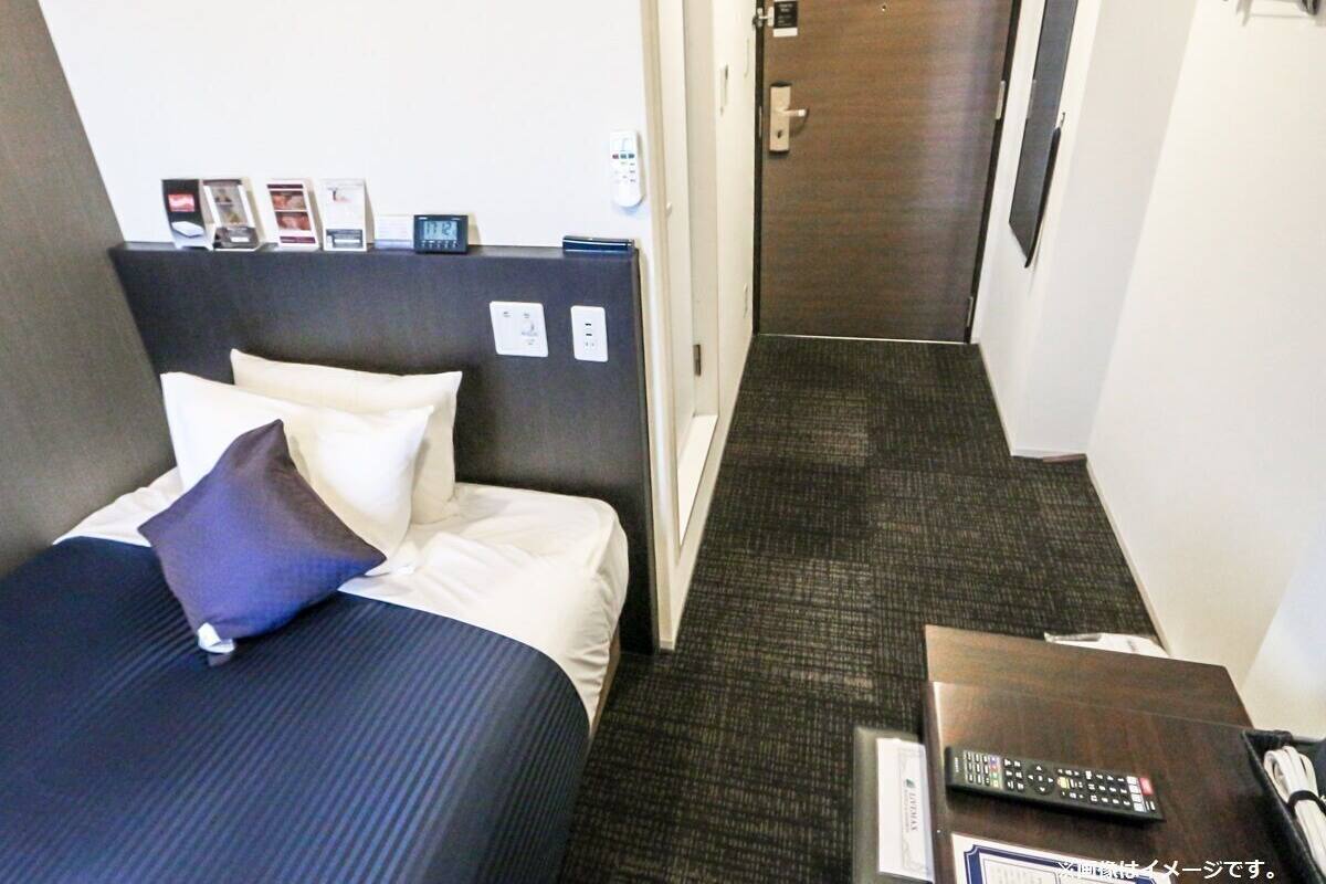 Quarto de hotel com cama, mesa de cabeceira com controle remoto, porta e relógio fixado na parede.