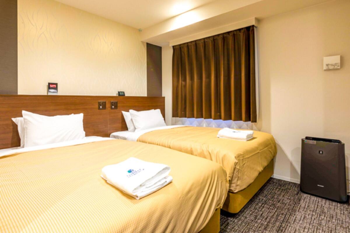Quarto de hotel com duas camas, cabeceira, janela com cortinas, escrivaninha e lixeira.