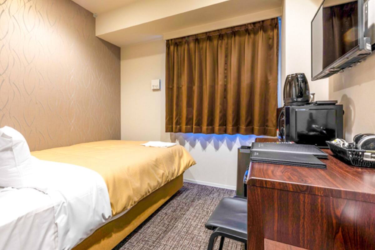 Quarto de hotel com cama, escrivaninha, cadeira, televisão e janela com cortinas.
