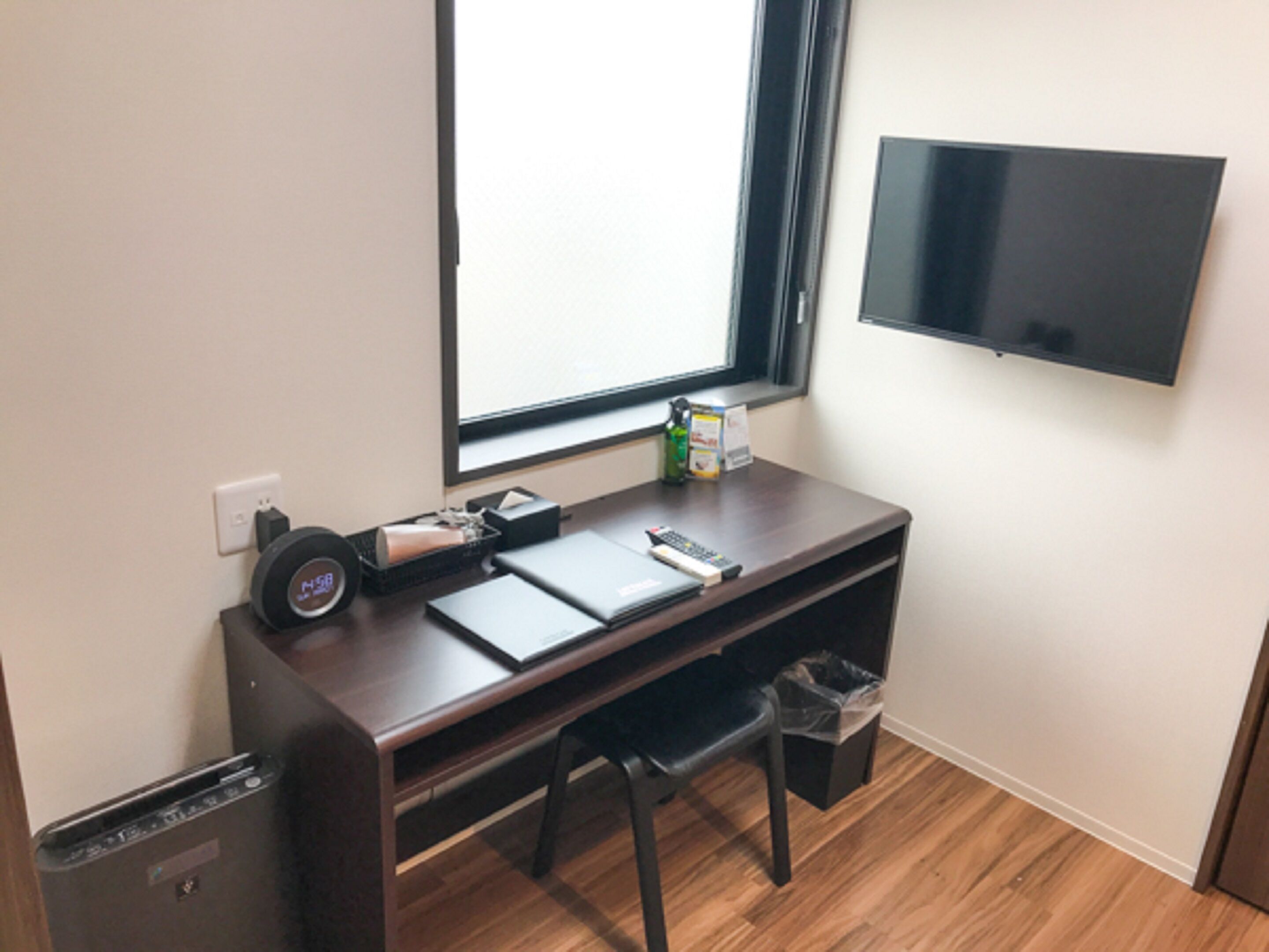 Una habitación pequeña con un escritorio de madera, una computadora portátil, un control remoto, un cesto de basura y un televisor montado en la pared.