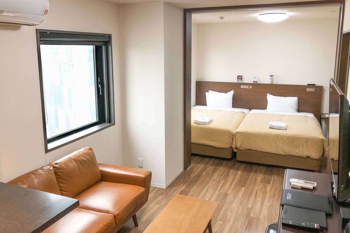 Ein Hotelzimmer mit braunem Ledersofa, einem Holztisch, zwei Betten mit beigefarbener Bettwäsche, einem Fenster und einem Fernseher auf einem Holzboden.