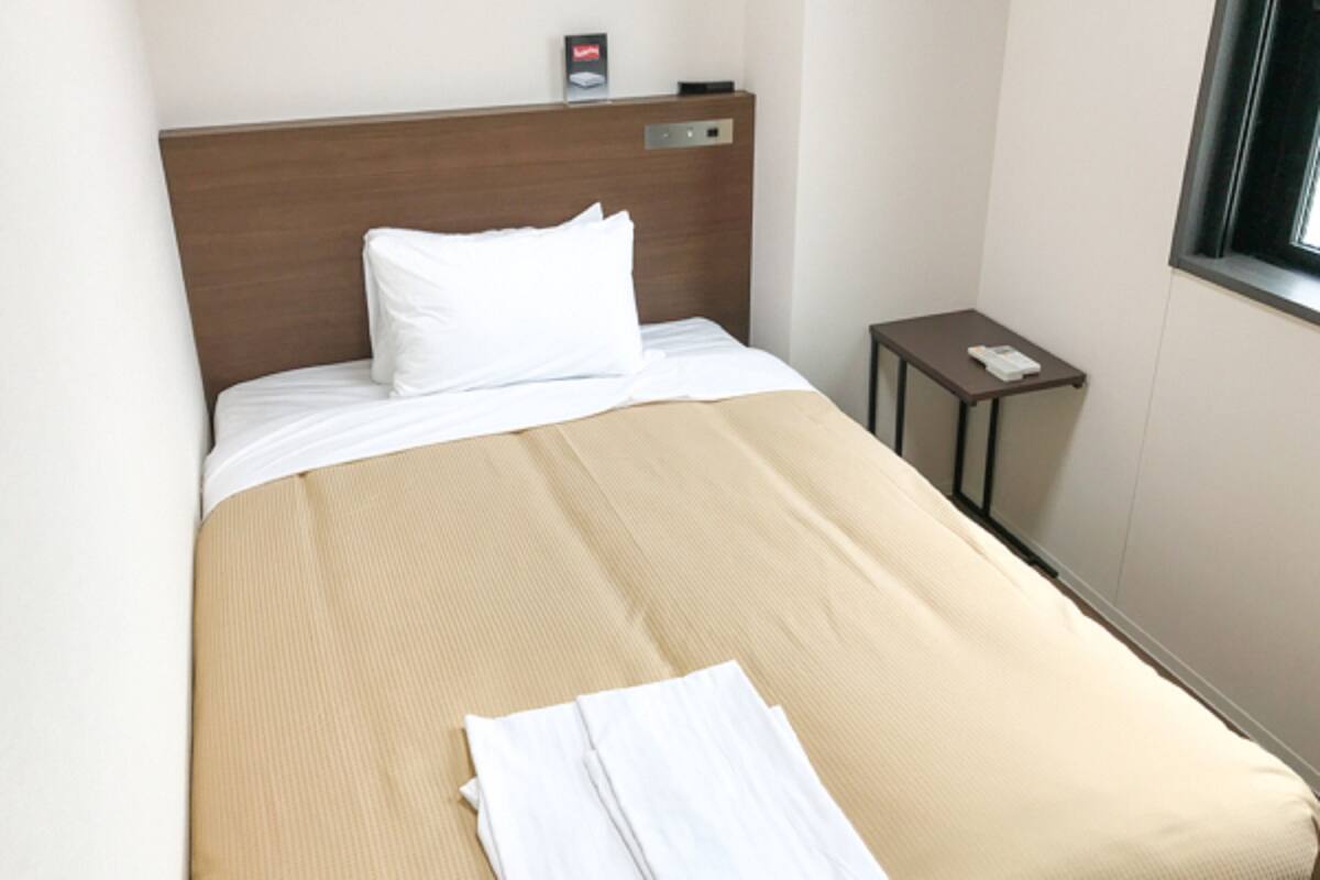 Ein ordentlich bezogenes Bett mit einer beigen Decke und weißen Kissen, ein kleiner Nachttisch mit einem Telefon und eine an der Wand montierte Bedienkonsole.