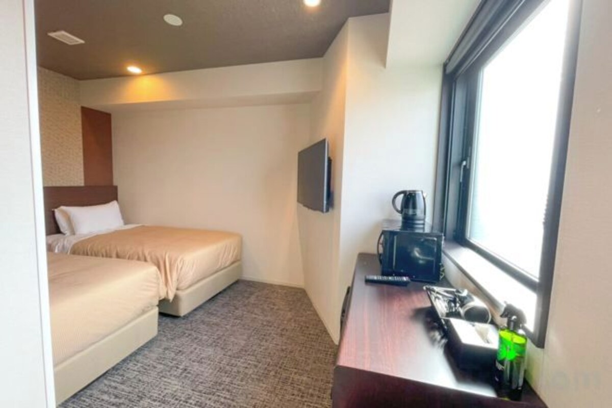 Quarto de hotel pequeno com uma cama, uma escrivaninha com cafeteira e uma televisão na parede.