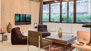 Ein modernes Wohnzimmer mit großem Fernseher, bequemen Sitzmöglichkeiten und Blick auf Grünfläche.