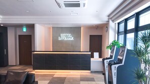 Un espace d’accueil d’hôtel au design moderne, comprenant un comptoir d’accueil, un coin salon avec un canapé et des fauteuils, et une grande fenêtre laissant entrer la lumière naturelle.