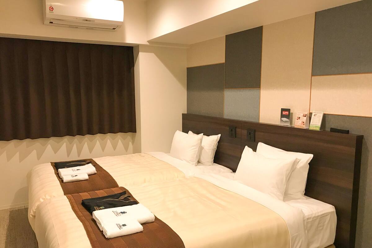 Ein Hotelzimmer mit einem großen Bett, einem Kopfteil mit integrierten Bedienelementen, einem Fenster mit Vorhängen und einer Wand-Klimaanlage.