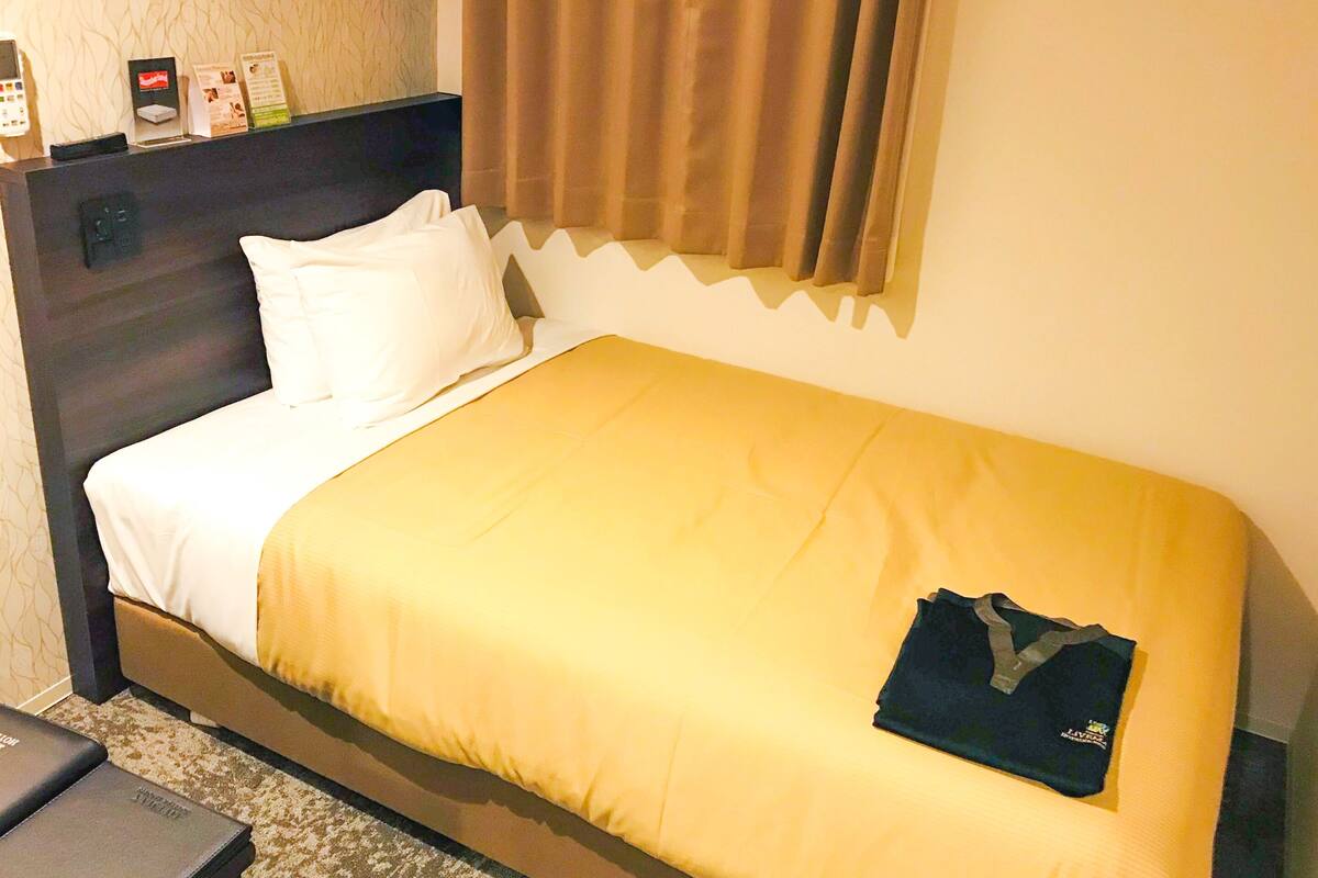 Ein Hotelzimmer mit Bett, Nachttisch, Kopfteil, Fenster mit Vorhängen und Wand mit strukturierter Oberfläche.