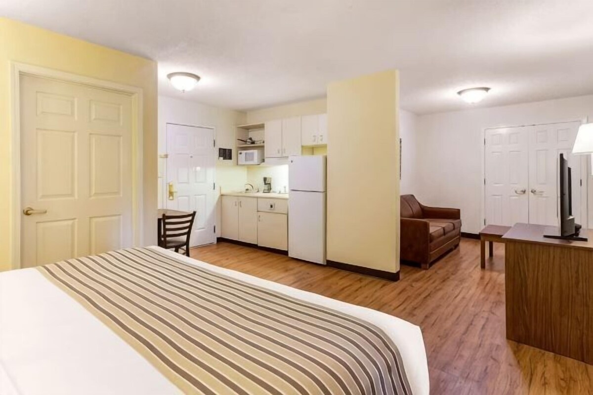 A compact hotel room with a bed, kitchenette, a small seating area, and a TV.