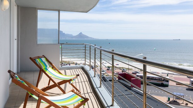 Ein Balkon mit zwei gestreiften Liegestühlen, Blick auf den Ozean, ein Berg in der Ferne und ein Parkplatz mit Bussen.