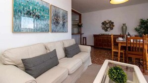A living room with a beige sofa, two abstract paintings, a dining area with a wooden table and chairs, and a hanging light fixture.