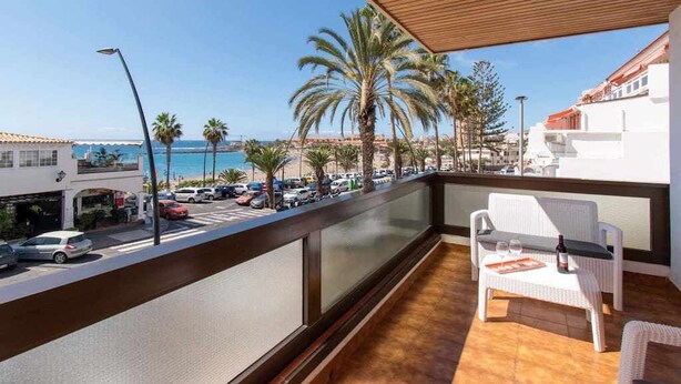 A balcony with a view of the sea, palm trees, and parked cars.