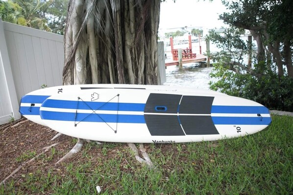 Une planche de surf blanche et bleue portant la marque « Vanhunks » et le numéro « 10 », posée sur l’herbe, près d’un arbre et d’une clôture blanche.