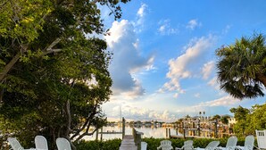 A waterfront view with a dock leading to a marina, surrounded by lush greenery and white outdoor seating. - Rare last-minute DISCOUNT in Indian Rocks Beach! Renovated 3BR WATERFRONT single family home with private dock sleeps 8, WALK to the sugar-white sand beach, Includes paddle board, beach chairs, wagon & umbrellas.  3bdrm, 2 bthrm, sleeps up to 8. (Indian Rocks Beach)