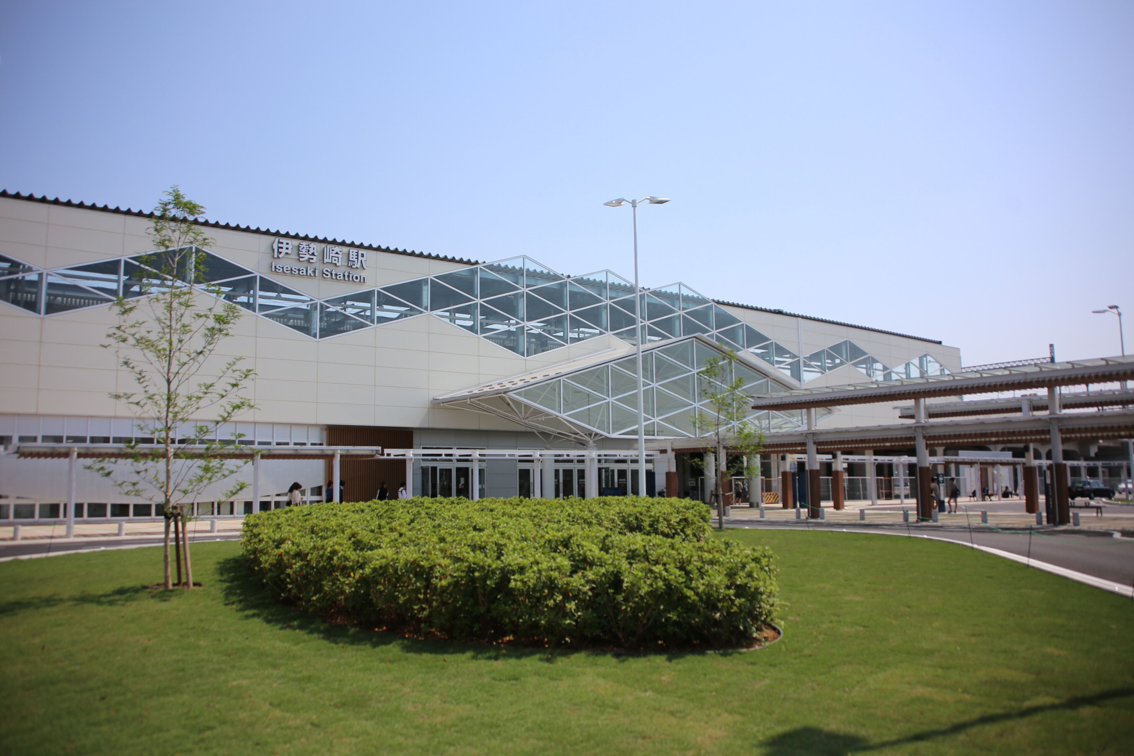 A modern building with a glass facade, a sign reading 'Ise Saki Station,' and a well-maintained lawn with a small tree.