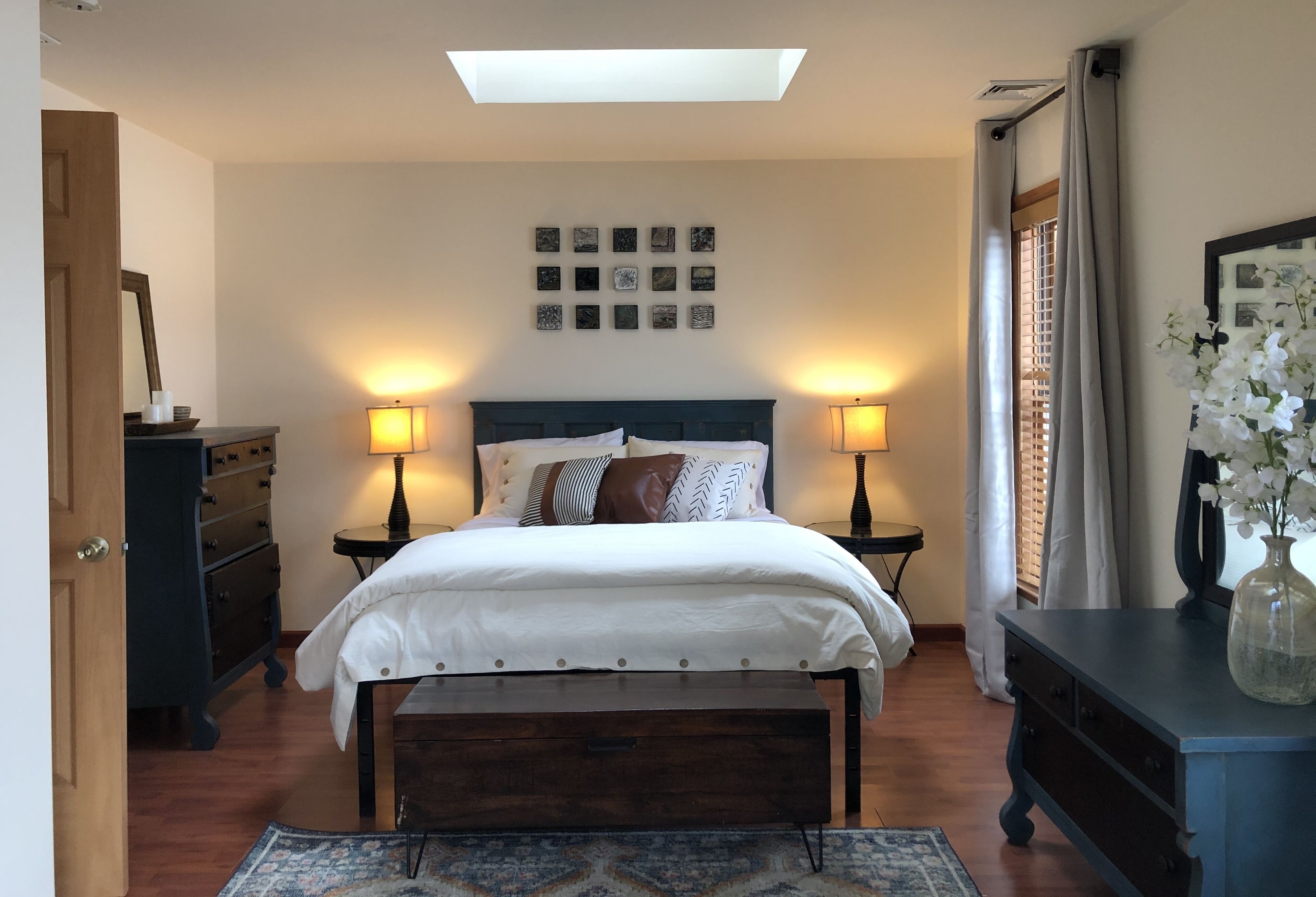 Un dormitorio con cama, lámparas de noche, cómoda, espejo, ventana con cortinas y tragaluz.