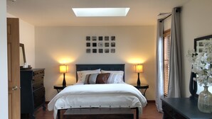 Un dormitorio con cama, lámparas de noche, cómoda, espejo, ventana con cortinas y tragaluz.