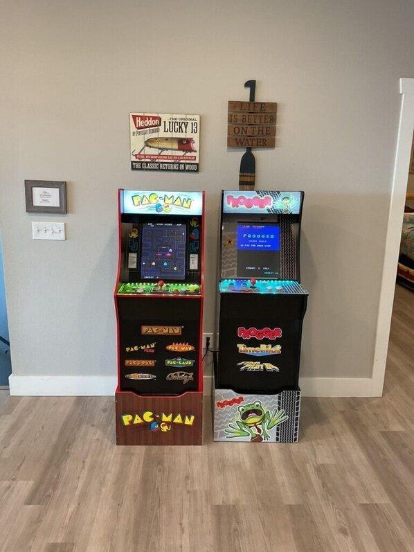 Dos máquinas de arcade, Pac-Man y Frogger, en una habitación con suelo de madera y un cartel en la pared que dice 'La vida es mejor en el agua'.