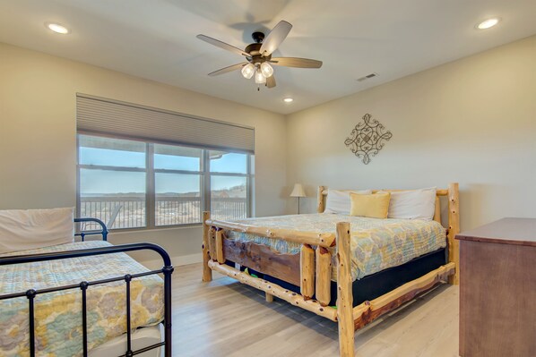 Un dormitorio con cama de madera, ventilador de techo, ventana grande y vista a la playa.