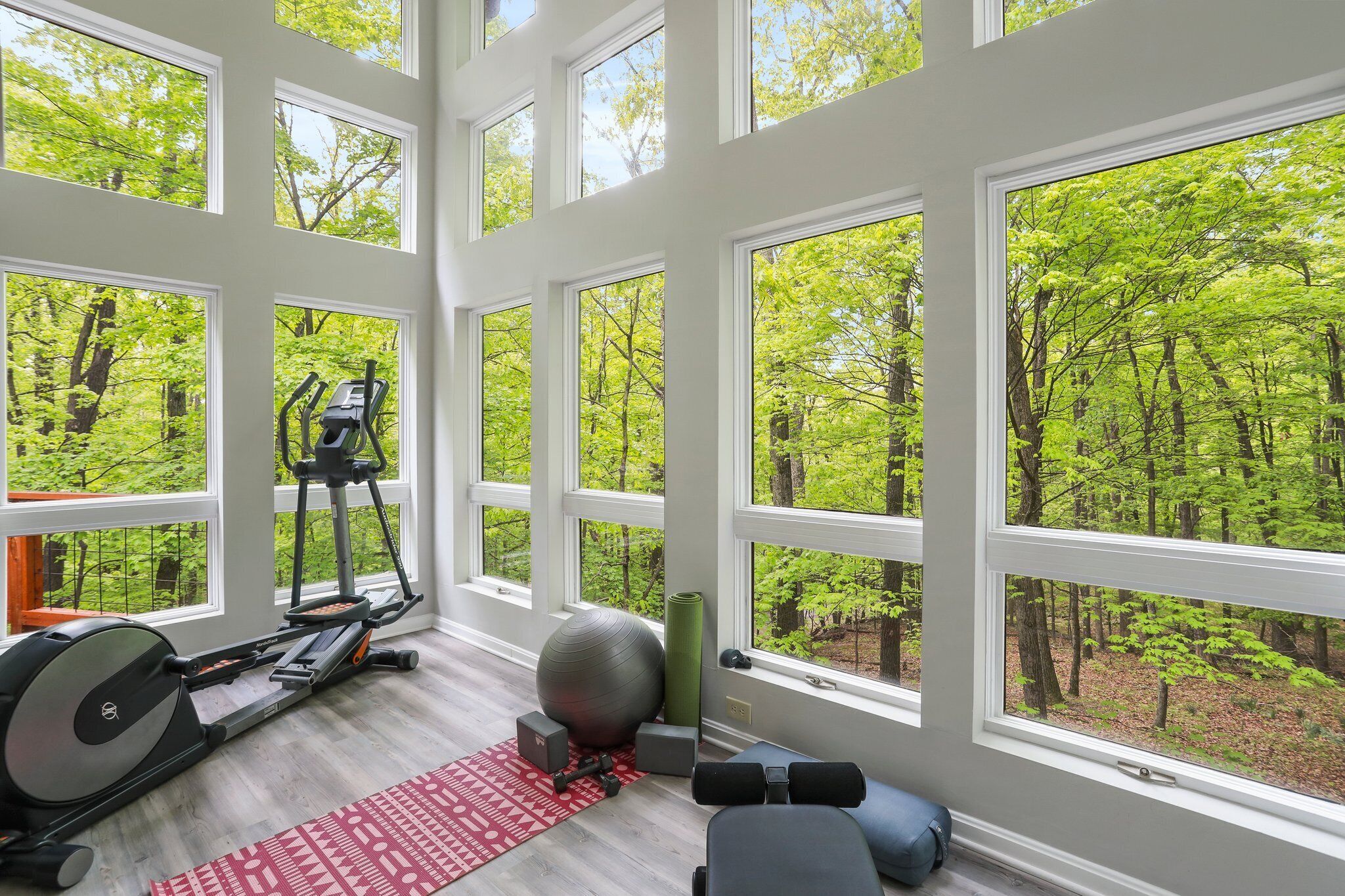 A gym with exercise equipment, a yoga mat, and large windows overlooking a forest.