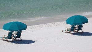 Zwei Strandstühle unter blauen Sonnenschirmen an einem sandigen Strand mit klarem, türkisfarbenem Wasser.