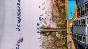 Vogelperspektive eines Strandes mit Reihen blauer und weißer Sonnenschirme, einem Pool und einem Gebäude mit Balkonen.