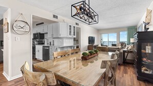 Eine moderne Küche mit weißen Schränken, ein Essbereich mit einem Glastisch und Holzstühlen sowie ein Wohnzimmer mit einer Couch und einem Kamin.