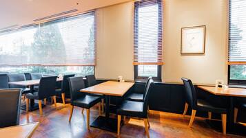 A restaurant with wooden tables and black chairs, large windows with blinds, and a framed picture on the wall.