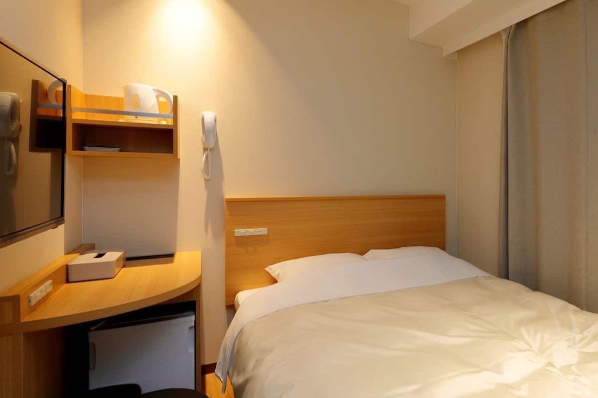 A hotel room with a bed, a desk with a telephone, a mirror, and a small shelf with toiletries.
